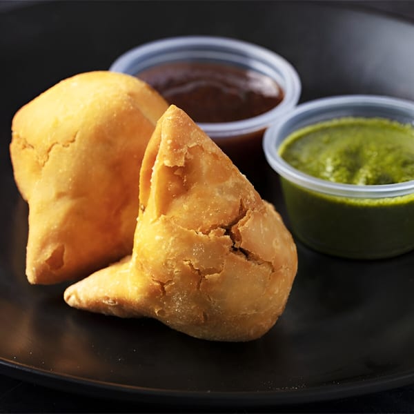 Two golden-brown samosas on a black plate, with green and brown chutneys in small plastic cups.